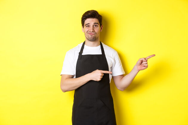 表情站在黄色背景上 戴着黑色围裙的可疑咖啡师右手指着手指 神情怀疑而不安工作室站立指点