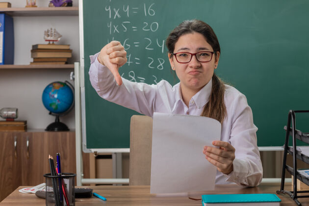 显示戴着眼镜的年轻女教师坐在课桌旁 空白的书页看起来困惑和不高兴 在教室的黑板前竖起大拇指检查作业女人检查老师