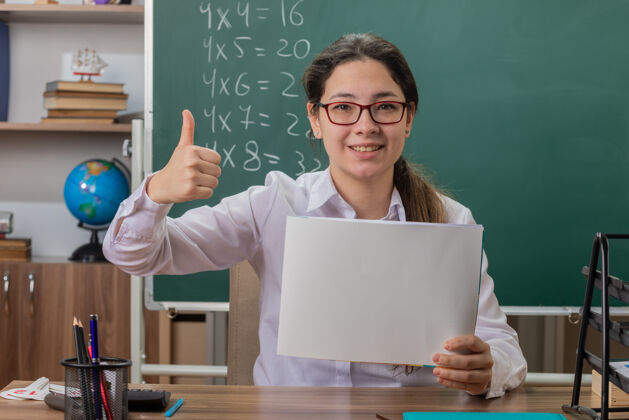 老师年轻的女老师戴着眼镜拿着空白的书页看着前面 兴高采烈地笑着竖起大拇指坐在教室黑板前的课桌旁拇指抱着展示