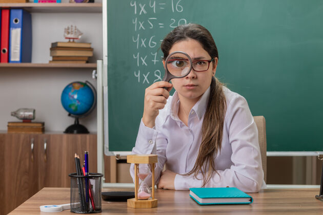 穿过年轻的女老师戴着眼镜 坐在教室黑板前的课桌上 透过放大镜看着前面眼镜前面玻璃