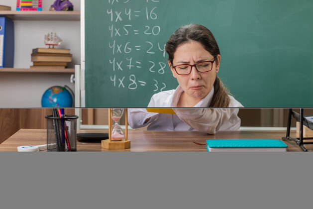 年轻人年轻的女老师戴着眼镜拿着书 用放大镜看着书页 自信的表情坐在教室黑板前的课桌旁桌子穿过自信