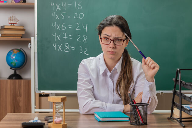 头年轻的女老师戴着眼镜拿着指针去讲课 看起来很困惑 坐在教室黑板前的课桌上挠头教室戴着黑板