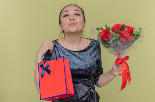 举行快乐的亚洲女人手持一束红玫瑰和一个带礼物的纸袋 站在绿色的墙上亲吻庆祝国际妇女节玫瑰庆祝吹