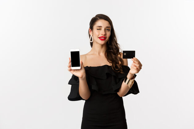 表情时尚购物理念红唇美女 对着相机眨眼 展示智能手机屏幕和信用卡 网购 白色背景魅力黑发莫比尔