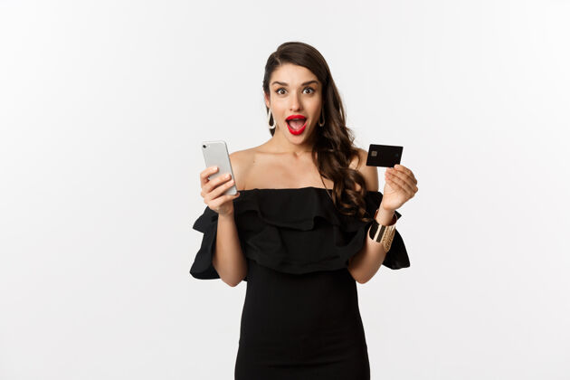 女性网上购物概念穿着黑色连衣裙的时髦女人 拿着智能手机的信用卡 看起来很兴奋 站在白色背景下网络营销黑发美丽