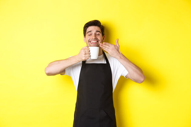 烹饪咖啡师坐在马克杯里享受着咖啡的味道 高兴地闭着眼睛站着 穿着黑色围裙自信黄色咖啡师