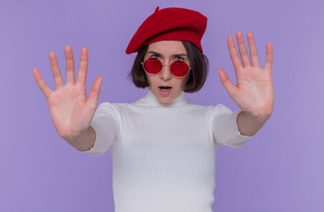 高领毛衣一个留着短发 头戴贝雷帽 头戴红色太阳镜的年轻女子 严肃地看着前面 做着停下来的手势 双手站在蓝色的墙上年轻穿手