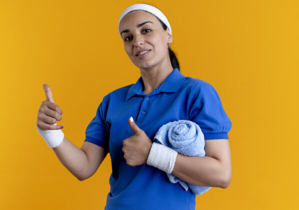 毛巾年轻自信的白人运动女性戴着头带和腕带大拇指向上拿着毛巾 手臂放在橙色的复制空间手臂橙色运动