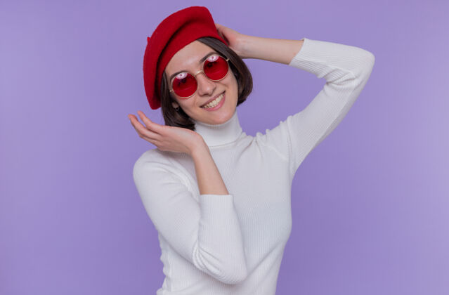 年轻快乐而积极的年轻女子 短发 白色高领毛衣 头戴贝雷帽 戴着红色太阳镜 站在蓝色的墙上 面带微笑 看着前方头发高领毛衣女人