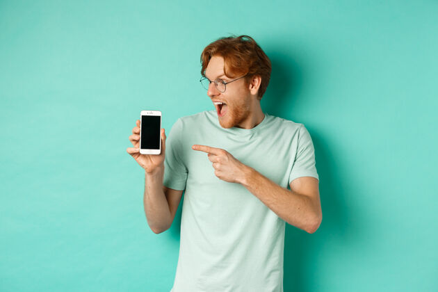 红发看看这个戴着眼镜的红发帅哥用手指着空白的智能手机屏幕 展示着网上的促销活动 站在绿松石色的背景下惊叹不已肖像屏幕显示