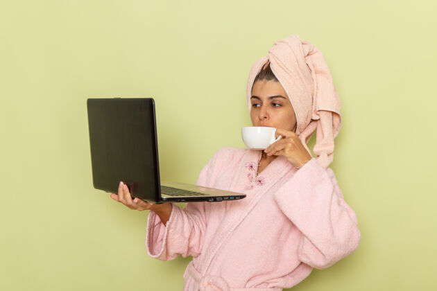 笔记本电脑前视图穿着粉色浴袍的年轻女性使用笔记本电脑 在绿色表面上喝咖啡咖啡自我保健年轻女性
