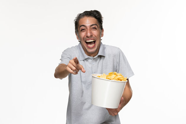 男正面图：年轻的男性正在吃薯片 在白色的表面大声大笑土豆前面大声