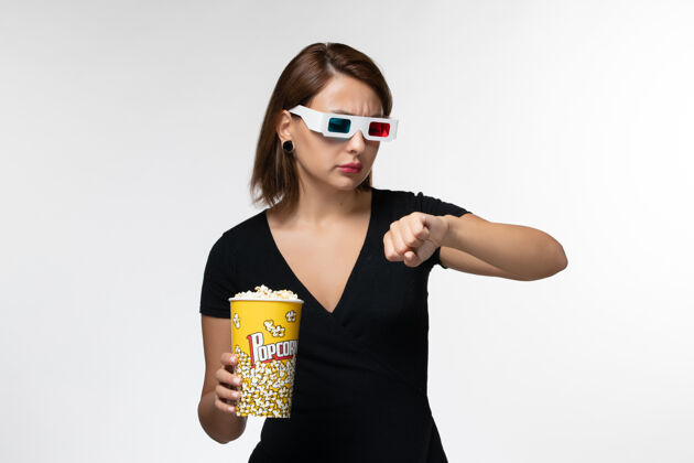电影正面图：戴着d型太阳镜拿着爆米花的年轻女性在白色表面上看着自己的手腕美丽年轻的女性手腕