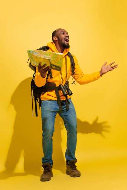 乐趣一个快乐的年轻游客的肖像 带着包和望远镜 隔离在黄色的工作室墙上地图光明人