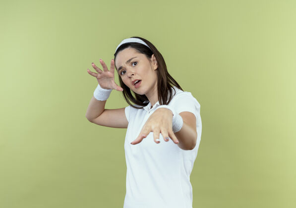 站立戴着头巾的年轻健身女士站在光墙上做着双手防御的姿势 脸上带着恐惧的表情表情女人头带