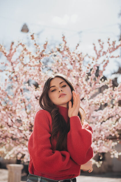 年轻在樱花的背景下 一个穿着鲜艳毛衣的漂亮女人看着镜头一张穿着红色毛衣的女人在外面摆姿势享受春天的快照贝雷帽花芳香