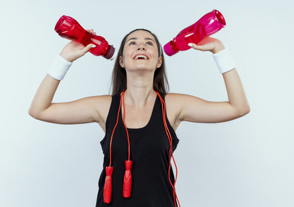 站着戴着头巾的年轻健身女 手拿一瓶水 站在白墙上 准备锻炼后喝水头带抱着奶瓶