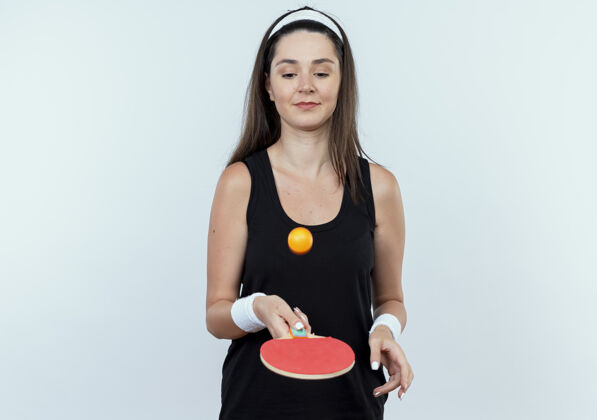 站着戴着头巾的年轻健身女士拿着球拍和乒乓球 微笑着自信地站在白色的墙上扔着球网球乒乓球年轻