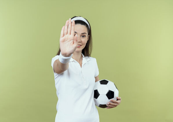 光戴着头巾 手持足球 手带微笑的年轻健身女士站在轻薄的墙上做停车标志女人立场健身