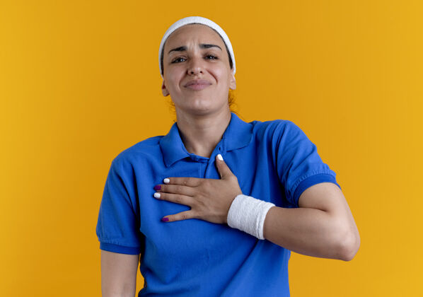 年轻年轻疼痛的高加索运动女性戴着头带和腕带把手放在胸部隔离在橙色空间与复制空间腕带复制手