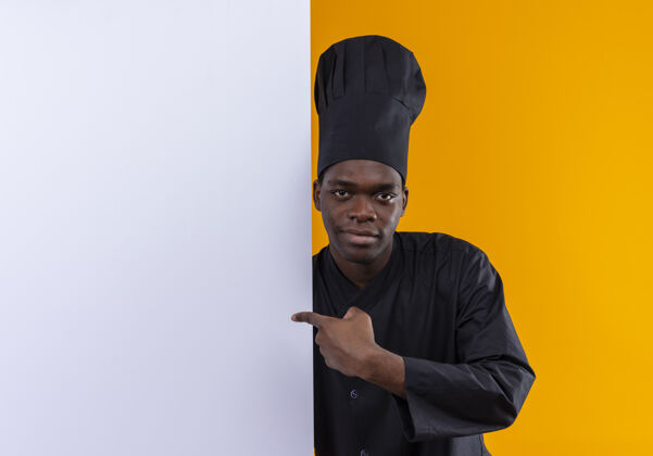 烹饪身着厨师制服的年轻自信的美国黑人厨师站在身后 指着隔离在橙色空间和复印空间的白色墙壁立场墙壁厨师