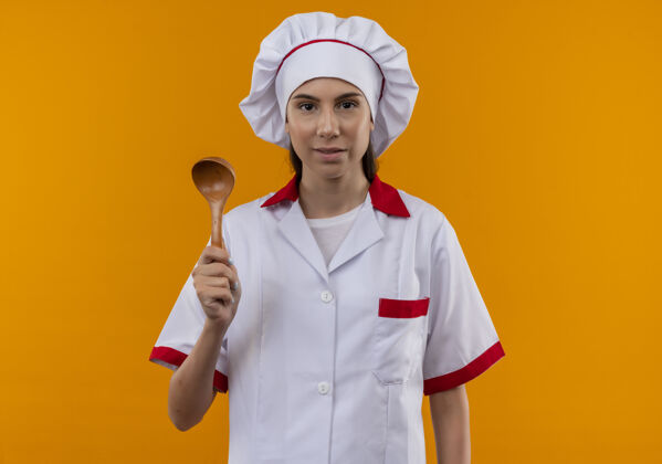 拿着穿着厨师制服的年轻快乐的白人厨师女孩拿着木勺子 在橙色的空间和复印空间隔离开来橙色请女孩
