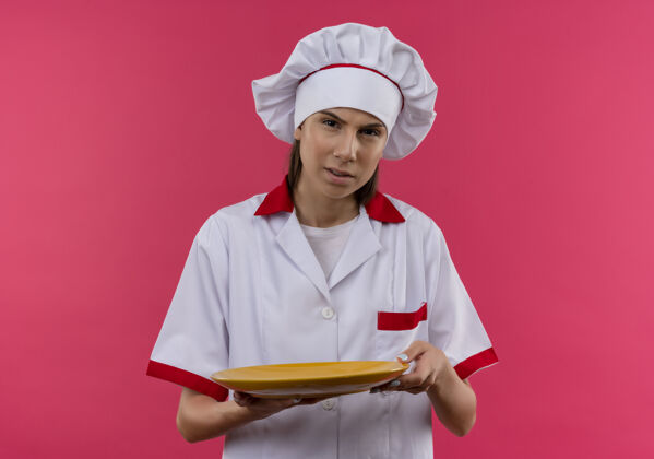 女孩穿着厨师制服的年轻恼怒的白人厨师女孩拿着盘子 把盘子孤立地放在粉红色的空间和复印空间里空间粉红年轻