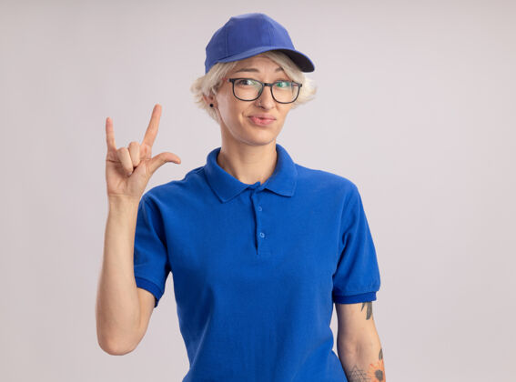 分娩身穿蓝色制服 戴着帽子的年轻女送货员看起来很困惑 站在白色的墙上做着摇滚的象征摇滚困惑女人