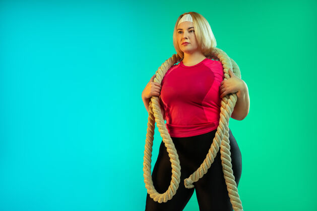 大小年轻的白人大号女模特在霓虹灯下的渐变绿色背景下训练用绳子做锻炼运动的概念 健康的生活方式 积极的身体 平等体重拉伸积极