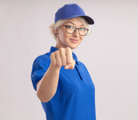 拳头身穿蓝色制服 头戴鸭舌帽的年轻女送货员站在白色的墙上 微笑着自信地站着交货制服微笑