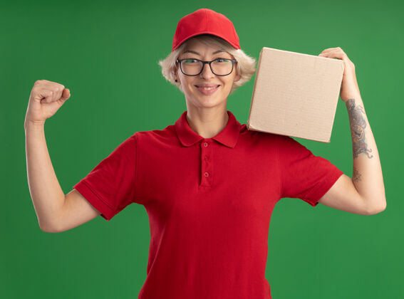 立场年轻的送货员身穿红色制服 戴着帽子 戴着眼镜 手里拿着纸板箱 举起拳头 微笑着 自信地站在绿色的墙上提高年轻信心