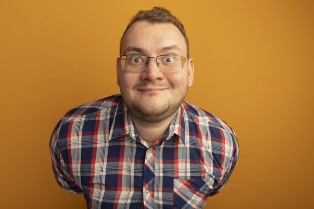 脸一个戴着眼镜和格子衬衫的男人站在橘色的墙上 面带微笑站着眼镜男人