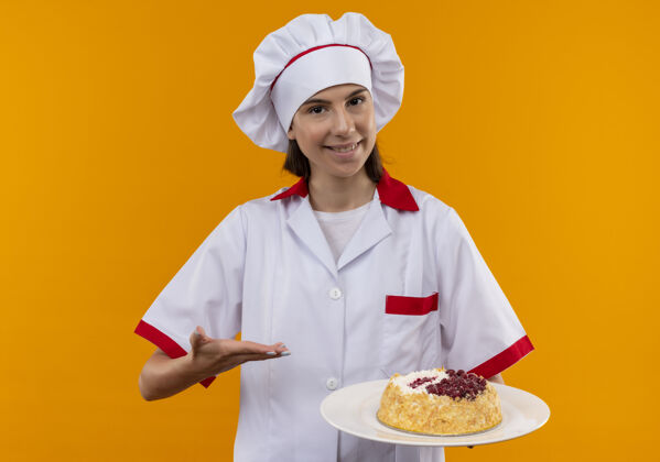 厨师身着厨师制服 面带微笑的年轻白人厨师女孩拿着并指着盘子上的蛋糕 蛋糕被隔离在橙色的空间里 还有复制空间空间年轻盘子