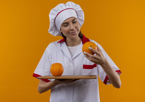 制服穿着厨师制服的年轻快乐的白人厨师女孩在砧板上拿着橘子 手上拿着橘子 在橘子空间和复印空间隔离开来请板女孩