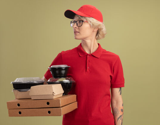 年轻人年轻的送货员身穿红色制服 戴着帽子 戴着眼镜 手里拿着披萨盒和食品包 严肃的脸站在绿色的墙上 望着一旁严肃戴着盒子