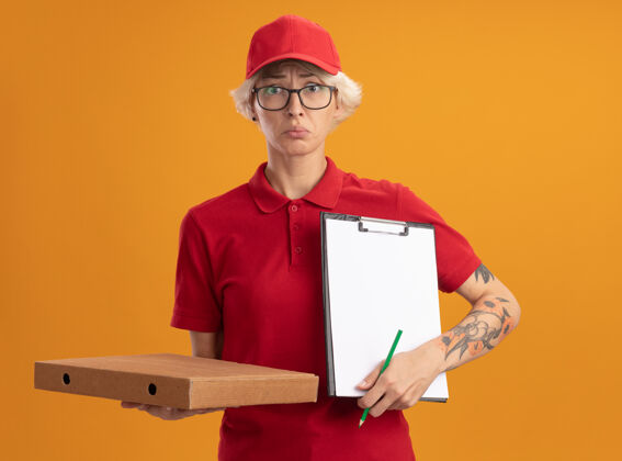 送货身穿红色制服 戴着帽子 戴着眼镜 拿着披萨盒和铅笔的剪贴板的年轻送货员站在橙色的墙上 神情迷茫 非常焦虑披萨帽子戴着