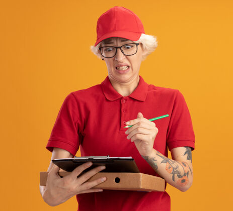 女人身穿红色制服 戴着帽子 戴着眼镜 拿着披萨盒和铅笔的剪贴板的年轻送货员站在橙色的墙上 神情困惑和失望站着拿着送货