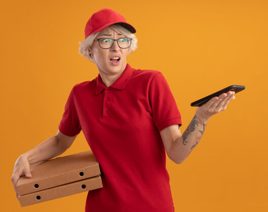 女士身穿红色制服 戴着眼镜 拿着比萨饼盒的年轻送货员站在橙色的墙上 看着自己的智能手机 困惑不已手机拿着困惑