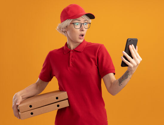 惊喜身穿红色制服 戴着帽子 戴着眼镜 拿着比萨饼盒的年轻送货员站在橙色的墙上 看着自己的智能手机屏幕 惊讶不已惊喜穿上屏风