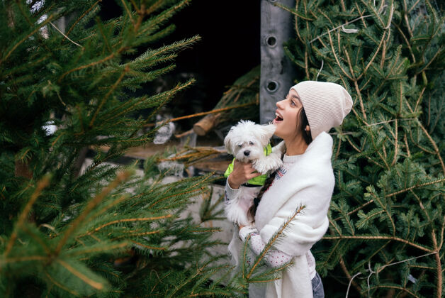 梗市场上一棵绿色的圣诞树旁 一个抱着一只白色狗的女人自然市场微笑