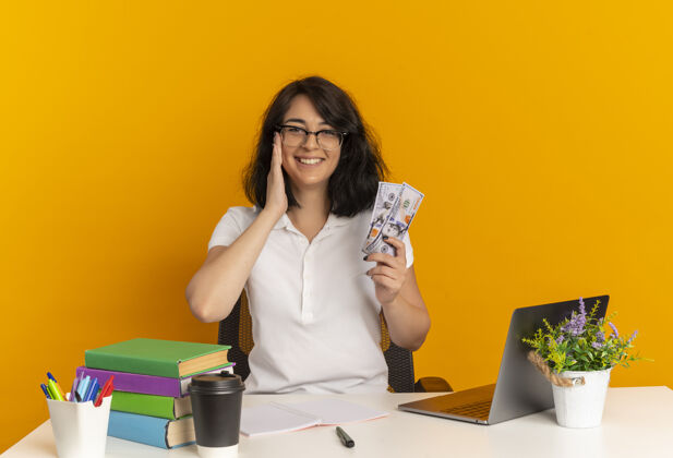 手年轻的笑容可掬的白人女学生戴着眼镜坐在书桌旁 手放在脸上 手里拿着钱隔离在橙色的空间和复印空间漂亮空间工具