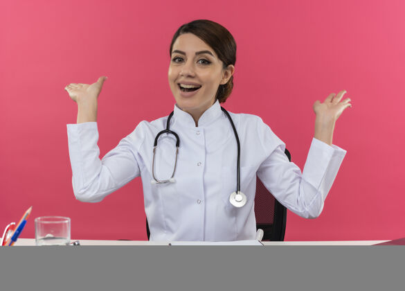 笔记本电脑快乐的年轻女医生穿着医用长袍和听诊器坐在办公桌旁 手里拿着医疗工具和笔记本电脑 两手空荡荡地站在粉红色的墙上女医疗办公桌