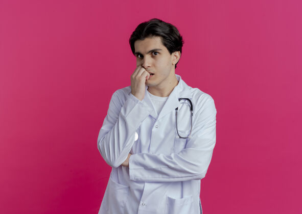 长袍焦急的年轻男医生穿着医用长袍和听诊器咬手指隔离在粉红色的墙上与复制空间看男性听诊器