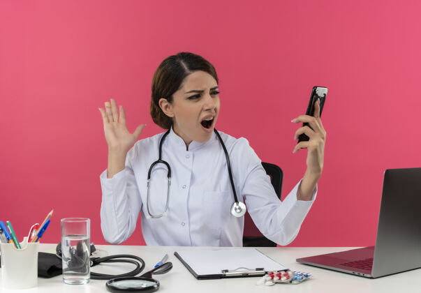 穿愤怒的年轻女医生穿着医用长袍和听诊器坐在办公桌旁 手里拿着医疗工具和笔记本电脑 手里拿着手机 手放在空气中 隔离在粉红色的墙上笔记本电脑医生手