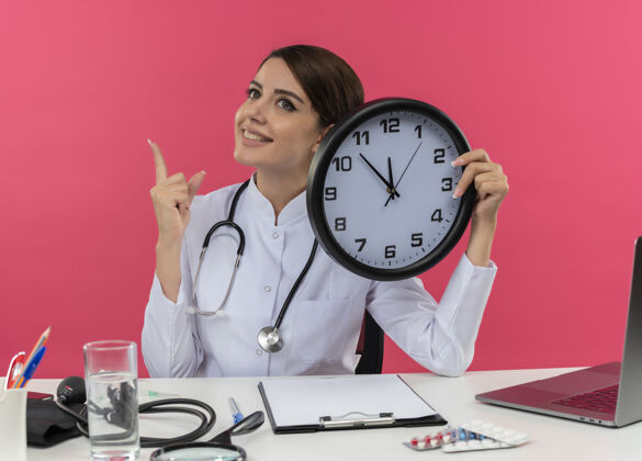 女面带微笑的年轻女医生穿着医用长袍 听诊器坐在办公桌旁 手里拿着医疗工具和笔记本电脑 手里拿着时钟 孤零零地看着粉红色的墙上时钟听诊器办公桌
