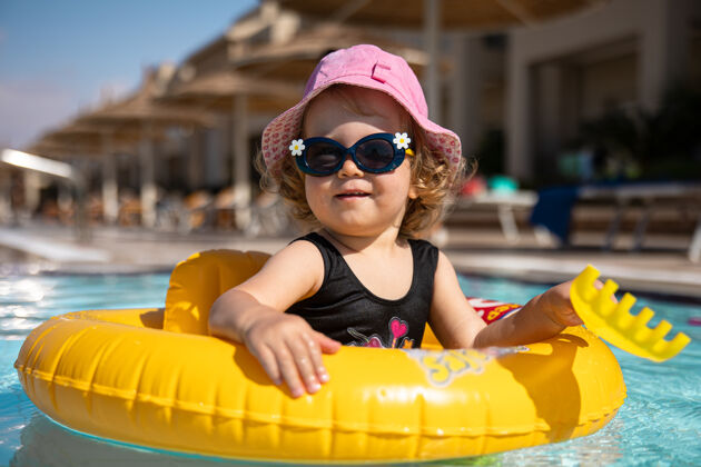 小戴着帽子和太阳镜的可爱小女孩坐在游泳池里玩游泳池阳光乐趣