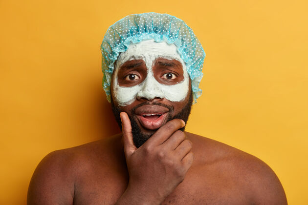 卫生非洲男人的头巾托着下巴 为了拥有健康的皮肤 涂上泥面膜 神情忧虑 戴上浴帽面膜完美化妆品