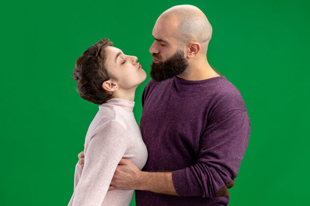 情侣一对穿着休闲装的年轻夫妇一个留着短头发的女人和一个留着胡须的男人在一起欢天喜地地拥抱着要亲吻庆祝情人节站在绿色的背景下拥抱在一起站立
