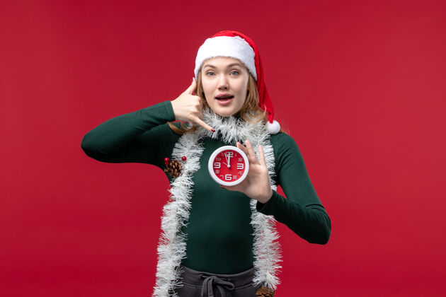 手持正面图红色背景上手持时钟的年轻女性成人圣诞节姿势