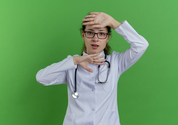 听诊器年轻的女医生穿着医用长袍 戴着听诊器和眼镜 手在头下和头上都被隔离在绿色的墙上穿看眼镜